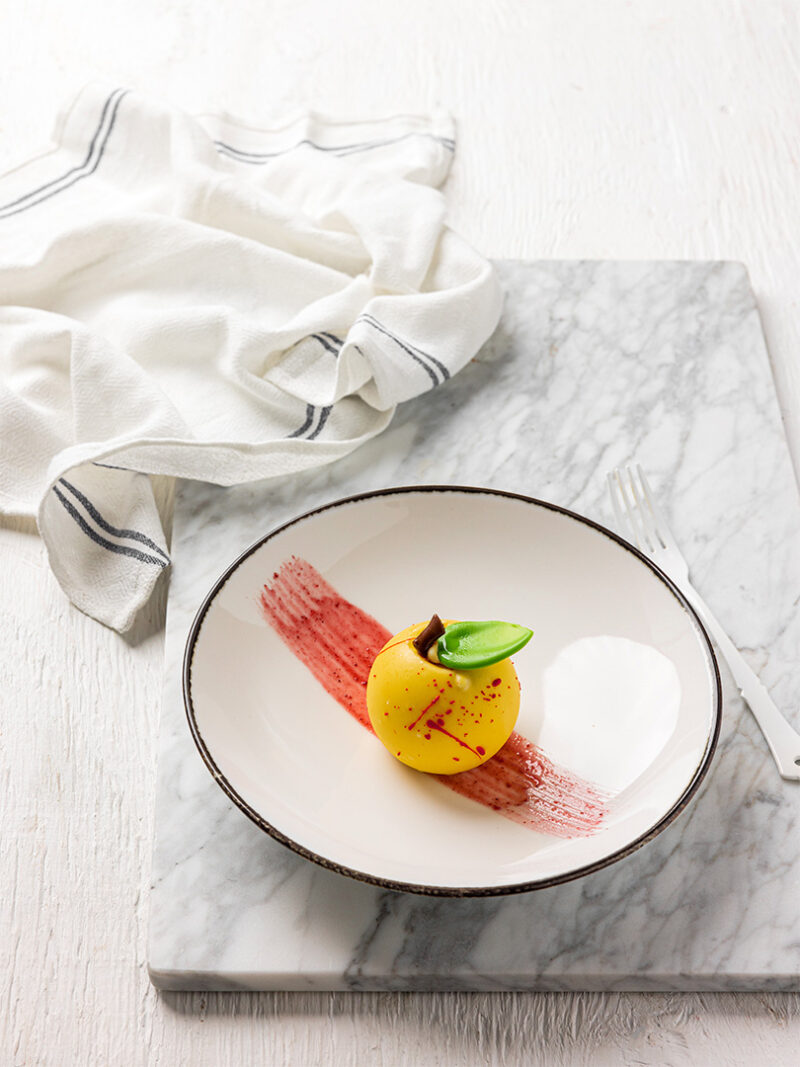 Dessert gastronomique en forme de pomme jaune avec feuille verte, dressé avec un trait de sauce rouge.