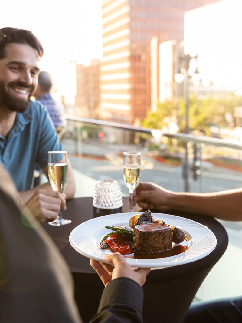 Assiette de filet de bœuf gourmet avec légumes servie sur une terrasse, deux personnes savourant du champagne en arrière-plan.