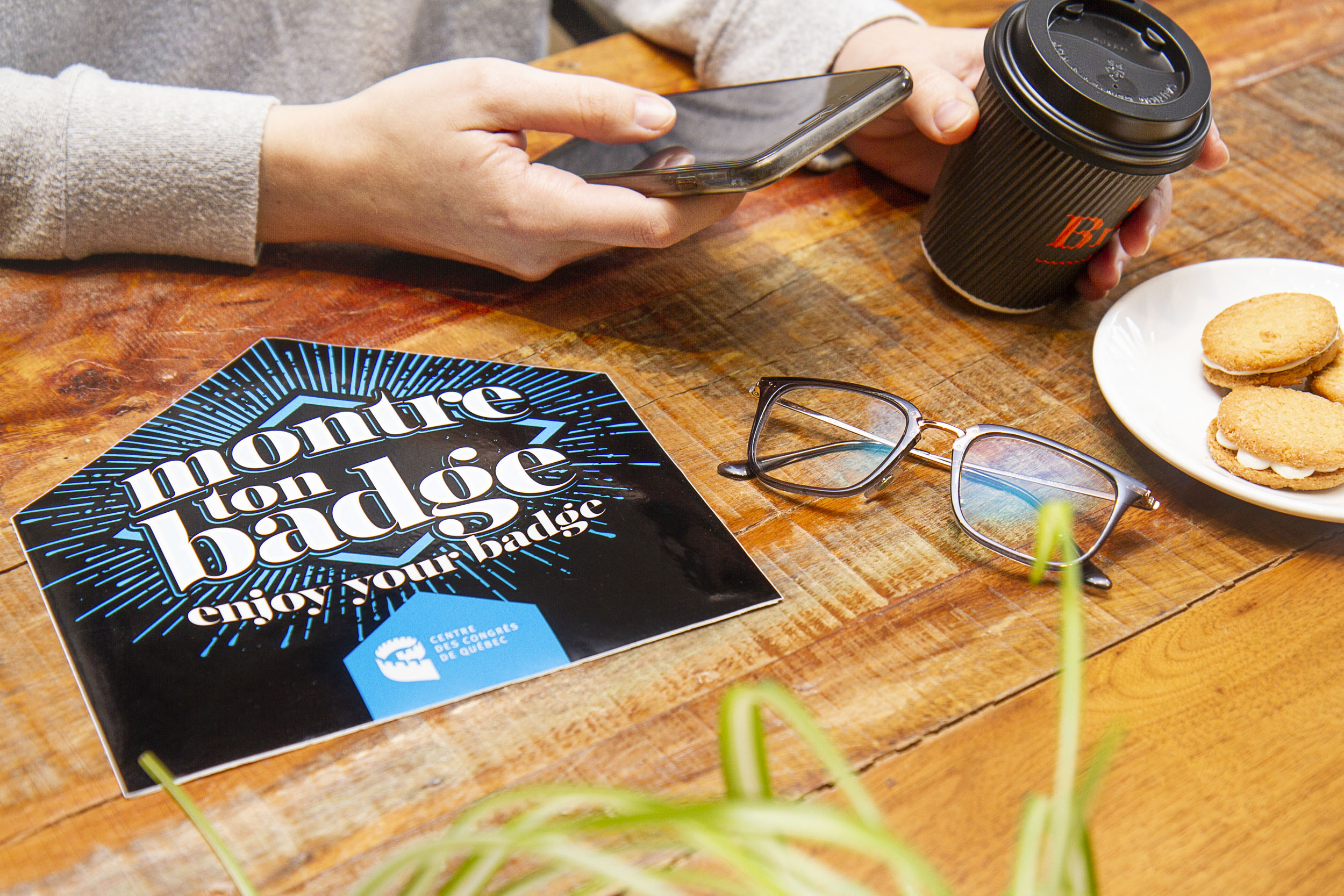 Carte “Montre ton badge” du Centre des congrès de Québec sur une table en bois, accompagnée d’un café à emporter, de lunettes et de biscuits pour profiter d’offres exclusives locales.